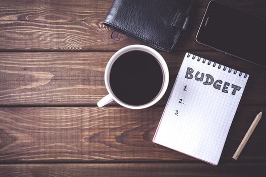 Top view of a white mug with black coffee next to a grid-lined notepad with the word "budget" at the top, a black leather wallet, and a black smartphone on a dark brown wood background.